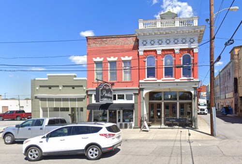 A street view of a historic building with a restaurant, featuring red brick and large windows, alongside a parked white SUV.