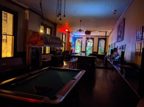 A dimly lit bar with a pool table, colorful lights, and empty seating areas.