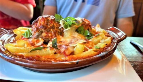 A close-up of baked pasta with meatballs, cheese, and fresh basil in a clay dish, served on a white plate.