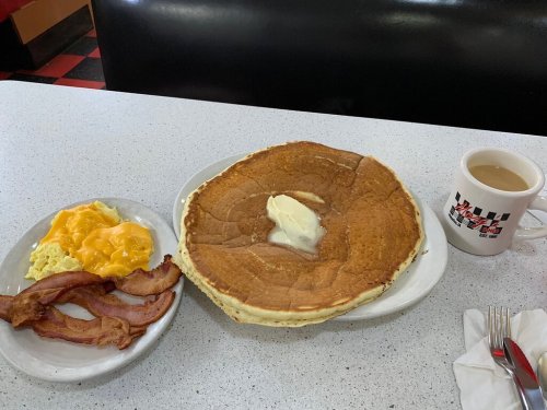 A large pancake with butter, scrambled eggs with cheese, bacon, and a cup of coffee on a diner table.