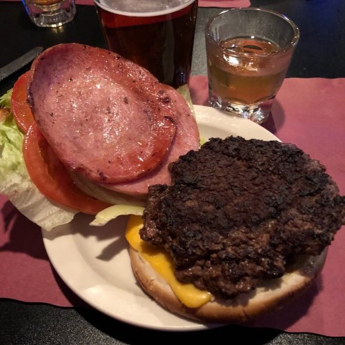 A plate with a burger topped with cheese, lettuce, tomato, and slices of ham, alongside a shot and a beer.