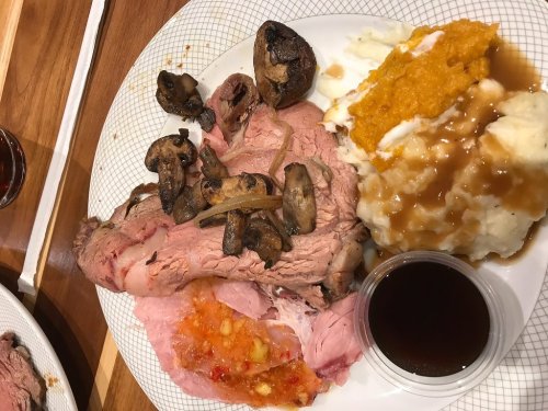 A plate of sliced roast beef with mushrooms, mashed potatoes, sweet potato, and a side of gravy.