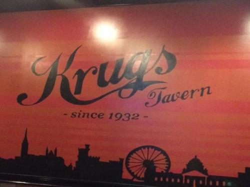 Sign for Krugs Tavern, featuring a vintage design with a sunset background and silhouettes of a city skyline.