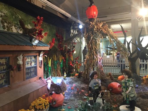 A festive display featuring a wooden cabin, pumpkins, autumn leaves, and animal figures in a seasonal setting.