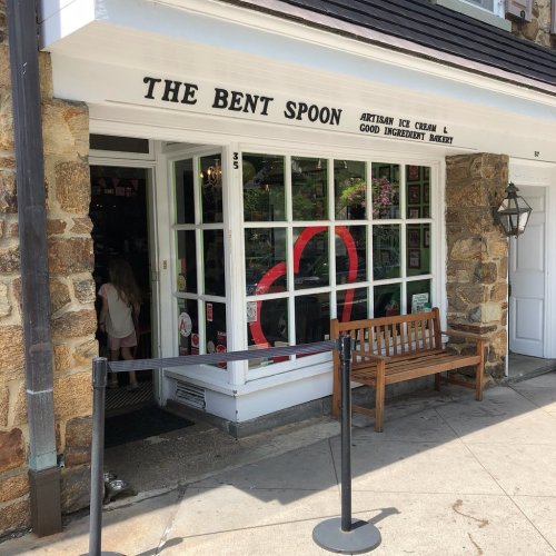 A quaint ice cream shop with a stone exterior and large windows, featuring a bench outside and a queue barrier.