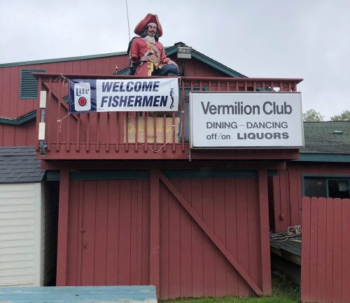 A large pirate figure sits atop a building with a sign welcoming fishermen and advertising the Vermilion Club.