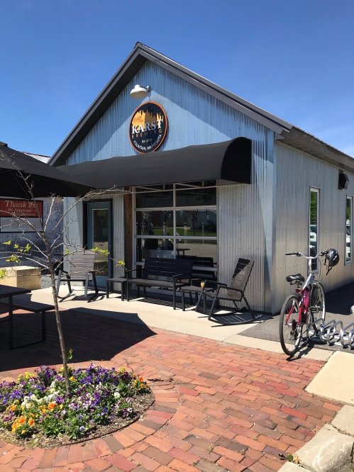 A modern café with a metal exterior, outdoor seating, and a bicycle parked nearby, surrounded by colorful flowers.