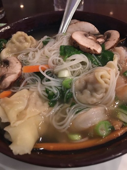 A bowl of soup with dumplings, mushrooms, vegetables, and rice noodles in a clear broth.