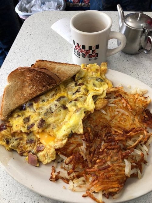 A plate with a cheesy omelet, crispy hash browns, and a slice of toast, alongside a coffee cup on a diner table.