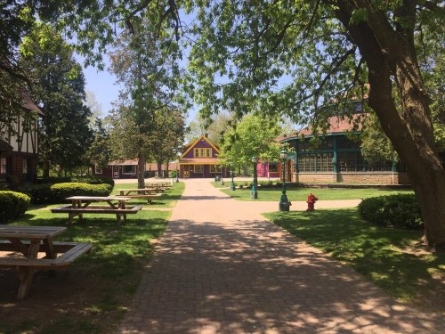 A sunny park pathway lined with trees, benches, and colorful buildings in the background.