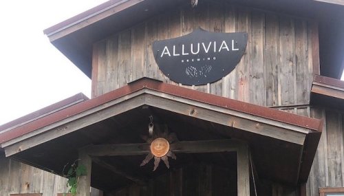Sign for Alluvial Brewing on a rustic wooden building with a sloped roof and decorative sun hanging below.