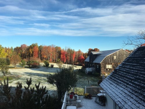 A scenic view of a farm with colorful autumn trees and a light dusting of snow on the ground and rooftops.