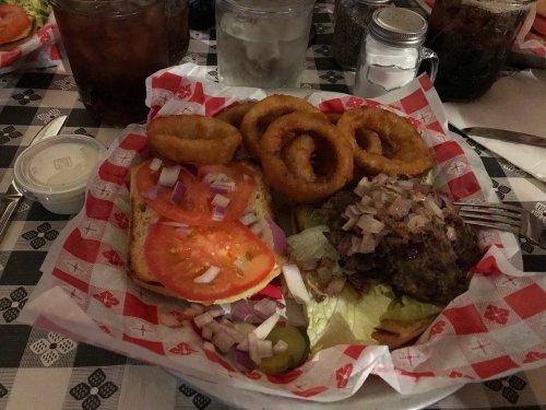 A plate featuring a burger with toppings, onion rings, and sides of lettuce, tomatoes, and pickles on a checkered tablecloth.