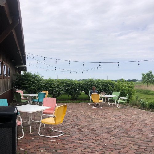 Outdoor seating area with colorful chairs and tables, string lights, and greenery under a cloudy sky.