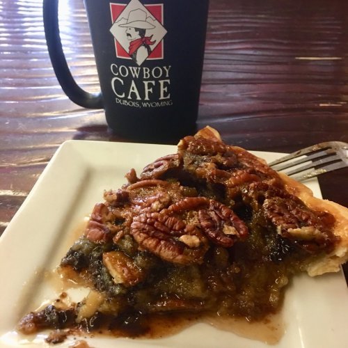 A slice of pecan pie on a white plate next to a black mug labeled "Cowboy Cafe, Dubois, Wyoming."