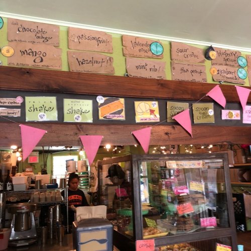 A cozy café interior with colorful menu boards, pastries, and a staff member behind the counter. Pink bunting decorates the space.