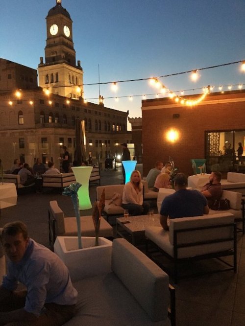 A rooftop lounge at dusk with string lights, modern seating, and a clock tower in the background. People are socializing.