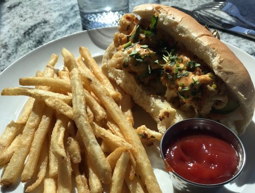 A plate with a sandwich filled with shrimp and herbs, served with a side of crispy fries and a small cup of ketchup.