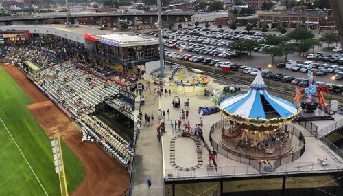 Aerial view of a baseball stadium with a carousel and carnival rides, surrounded by fans and parked cars.