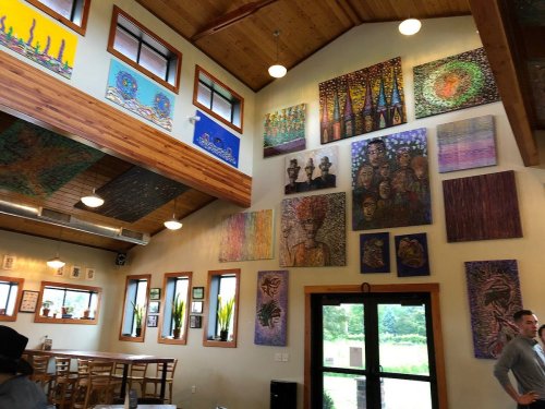 Interior of a cozy café featuring colorful artwork on the walls and wooden furniture, with large windows letting in light.