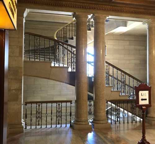 A grand staircase with ornate railings, flanked by columns, in a well-lit, elegant interior space.