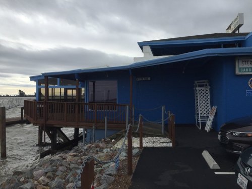 A blue waterfront building with a deck, surrounded by rocks and water, under a cloudy sky.