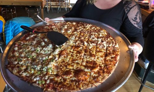 A woman holds a large pizza on a tray, smiling in a cozy restaurant with wooden decor.