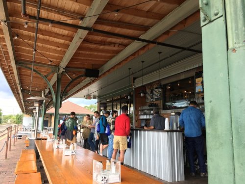 A busy outdoor café with people waiting in line and seated at long wooden tables under a covered patio.