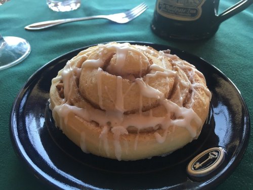 A freshly baked cinnamon roll drizzled with icing, served on a black plate with a green tablecloth in the background.