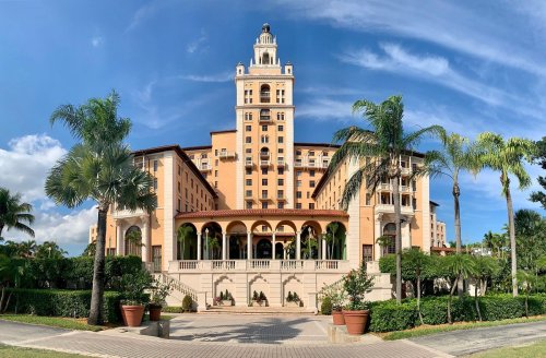 A grand hotel with a tall tower, surrounded by palm trees and lush greenery under a blue sky.
