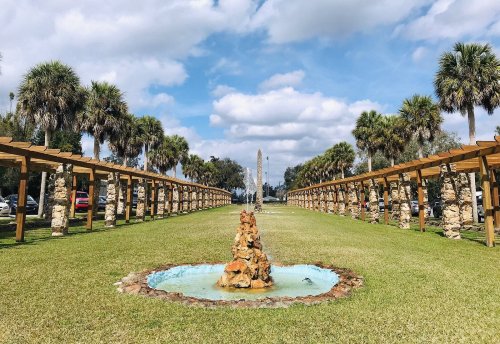 A landscaped park featuring a stone fountain, palm trees, and a pathway lined with wooden structures under a blue sky.