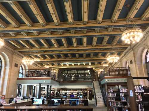 Interior of a library featuring ornate ceiling, bookshelves, and chandeliers, with study areas and a staircase visible.