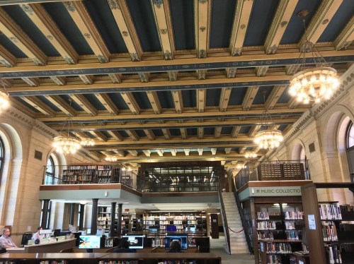 Interior of a library featuring ornate ceiling, bookshelves, and chandeliers, with study areas and a staircase visible.