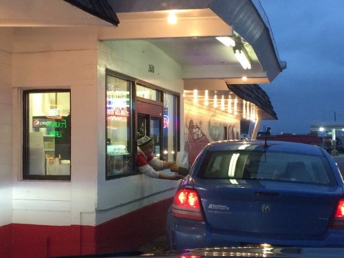 A drive-thru worker hands a bag to a customer in a blue car at dusk, with neon lights in the background.
