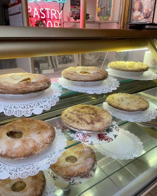 A display case filled with various pies on decorative doilies, with a neon pastry shop sign in the background.