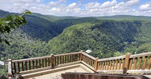 A scenic view of lush green mountains and a river valley under a partly cloudy sky from a wooden overlook.