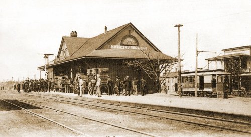Historic black-and-white photo of a train station with people waiting and vintage trains nearby.