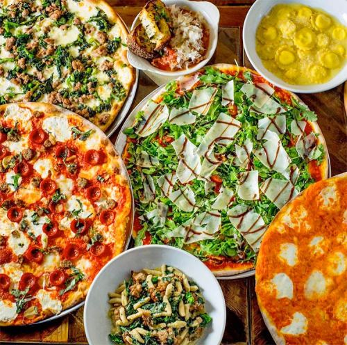 A variety of pizzas and pasta dishes arranged on a wooden table, featuring toppings like pepperoni, greens, and cheese.