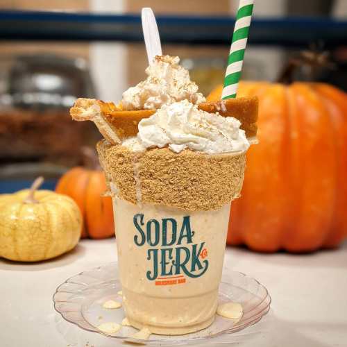 A decadent milkshake topped with whipped cream, graham cracker crumbs, and a slice of pie, with a pumpkin in the background.