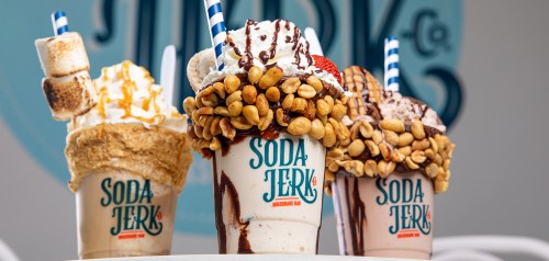 Three colorful milkshakes topped with whipped cream, nuts, and marshmallows, served in branded cups from Soda Jerk.