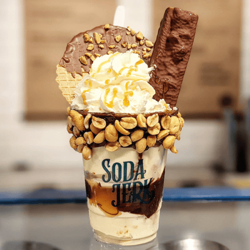 A decadent milkshake topped with whipped cream, chocolate, a cookie, and crushed peanuts in a branded cup.