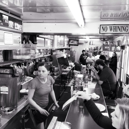 A busy diner scene in black and white, featuring a waitress serving customers at the counter and people dining.