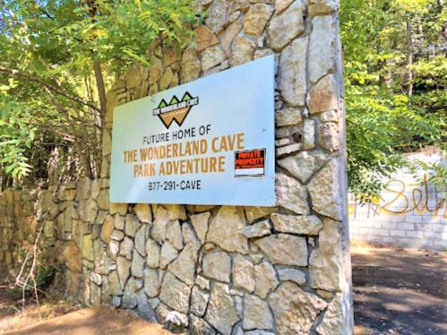 Sign for the future home of Wonderland Cave Park Adventure, surrounded by trees and stone wall.