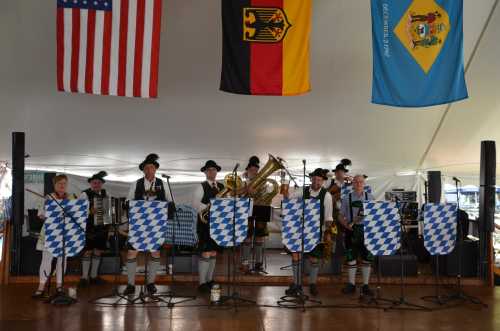 A band in traditional attire performs under flags, with instruments and blue and white shields in front of them.