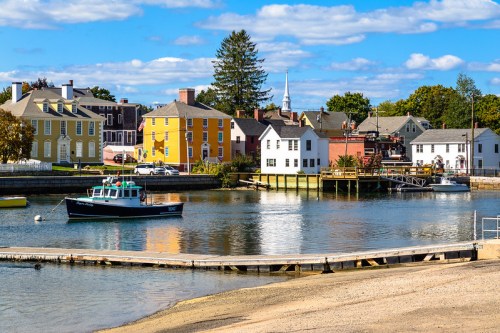 A scenic waterfront view featuring colorful houses, a boat, and a calm river under a blue sky with fluffy clouds.