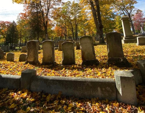 Gravestones in a cemetery surrounded by colorful autumn leaves and trees.