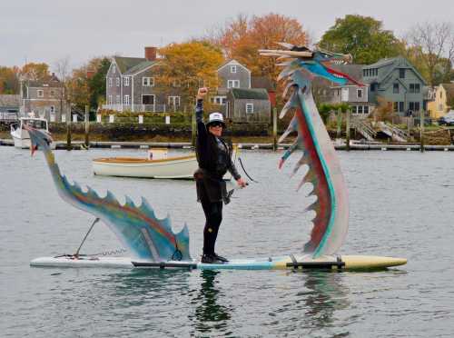 A person stands on a colorful dragon-shaped paddleboard in a harbor, raising a fist in celebration.