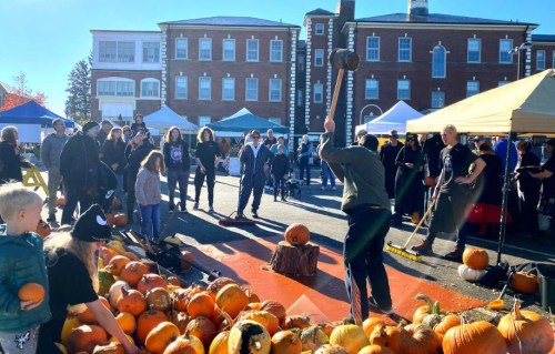 A crowd watches as a person swings an axe over a pile of pumpkins at an outdoor event.