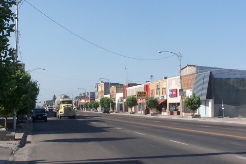 A quiet street lined with shops and trees, featuring a truck and clear blue sky in the background.