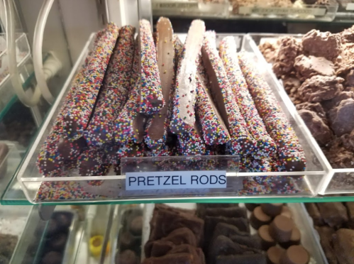 A display of pretzel rods coated in chocolate and colorful sprinkles, arranged neatly in a clear container.